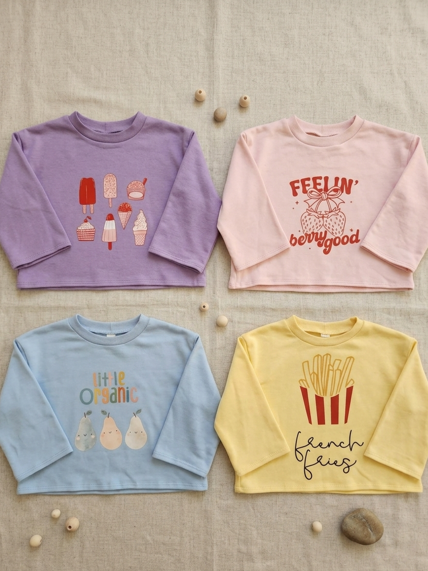 Four children's sweatshirts in purple, pink, blue, and yellow with various prints on a textured surface.