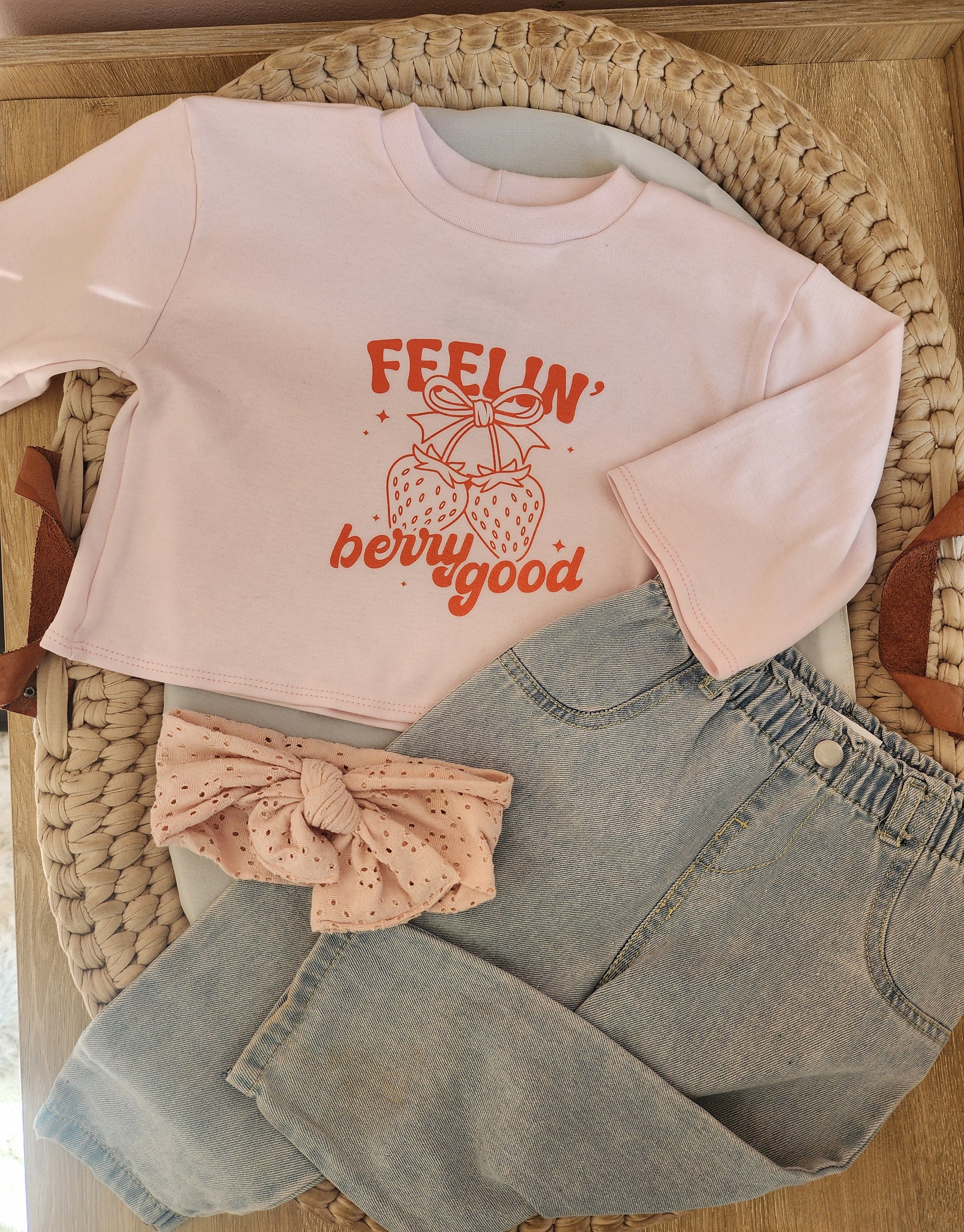 Pink long-sleeve shirt with 'FEELIN' berry good' text, denim shorts, and pink headband on a woven surface.