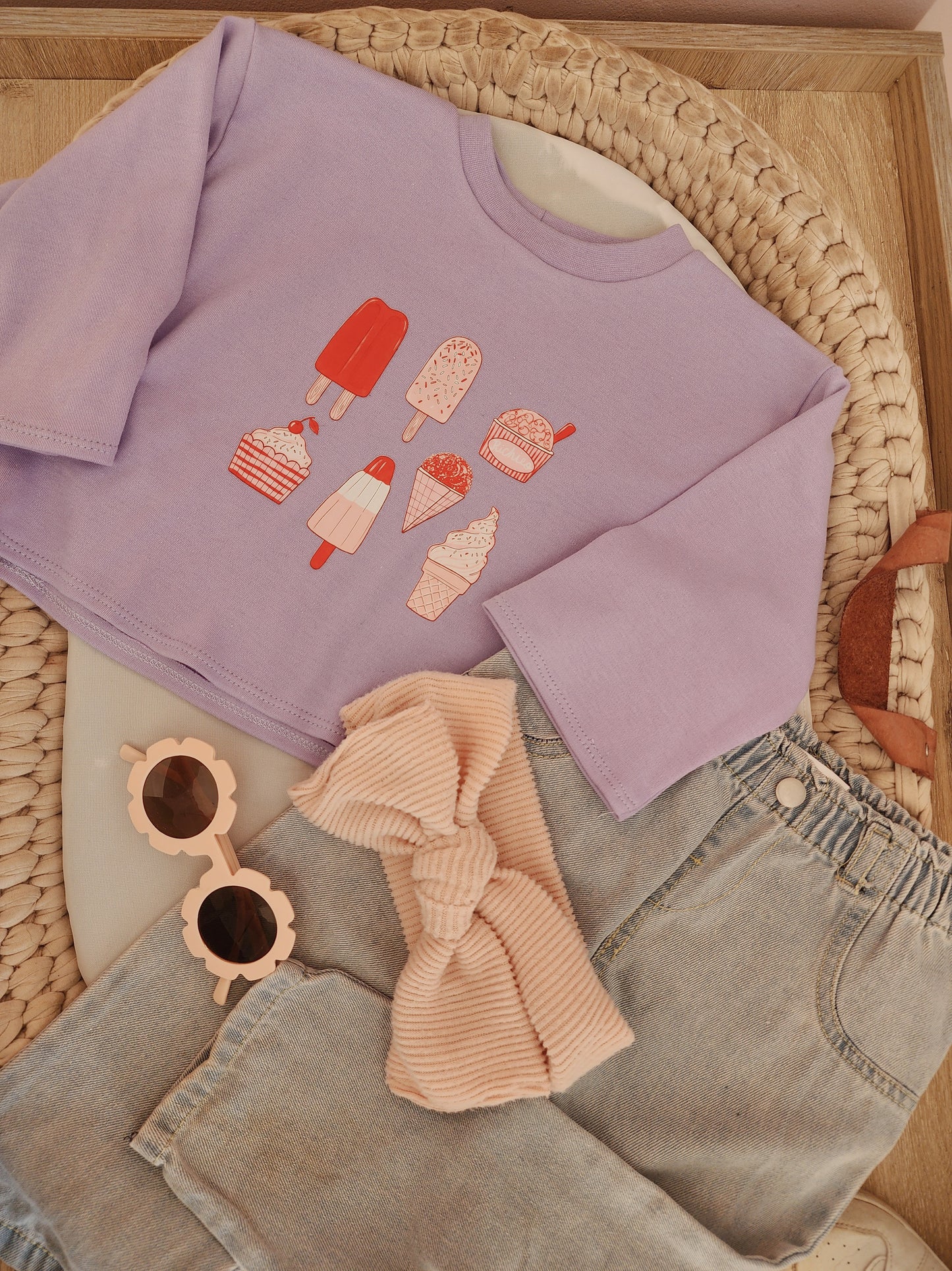 Purple sweatshirt with ice cream graphics on a woven surface with sunglasses and jeans.