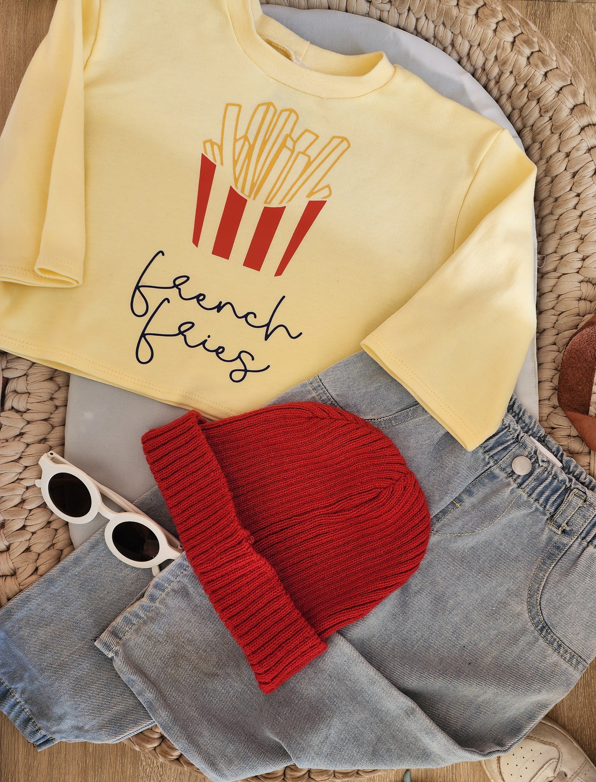 Yellow sweatshirt with 'French Fries' design, red beanie, sunglasses, and denim shorts on a woven surface.